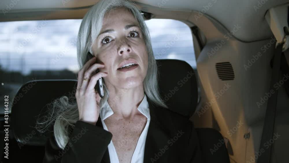 Woman in official suit talking on smartphone while driving in auto. Caucasian middle-aged gray-haired businesswoman holding laptop on knee, calling to business partner. Communication concept. Woman in official suit talking on smartphone while driving in auto. Caucasian middle-aged gray-haired businesswoman holding laptop on knee, calling to business partner. Communication concept.