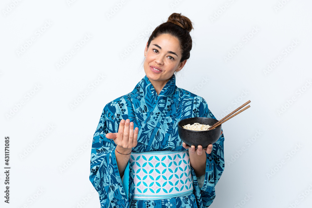 Woman wearing kimono over isolated white background inviting to come with hand. Happy that you came while holding a bowl of noodles with chopsticks Woman wearing kimono over isolated white background inviting to come with hand. Happy that you came while holding a bowl of noodles with chopsticks