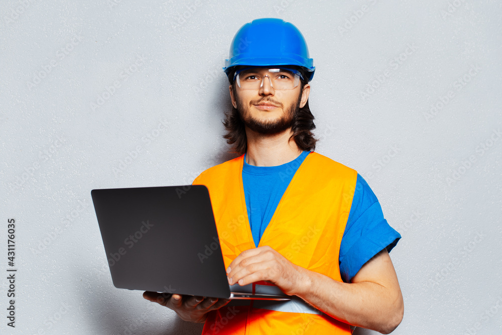 Portrait of young confident man, construction worker engineer wearing safety equipment, using laptop on background of grey wall. Portrait of young confident man, construction worker engineer wearing safety equipment, using laptop on background of grey wall.