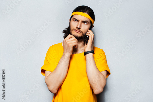 Studio portrait of young thoughtful man talking on smartphone, looking up, against background of grey wall. Guy wearing yellow shirt and hair band.