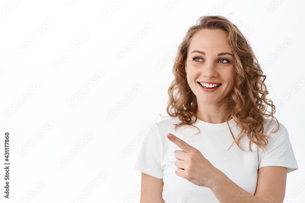 Young caucasian woman with fair hair, smiles and points aside at copyspace, shows advertisement, stands over white background Young caucasian woman with fair hair, smiles and points aside at copyspace, shows advertisement, stands over white background