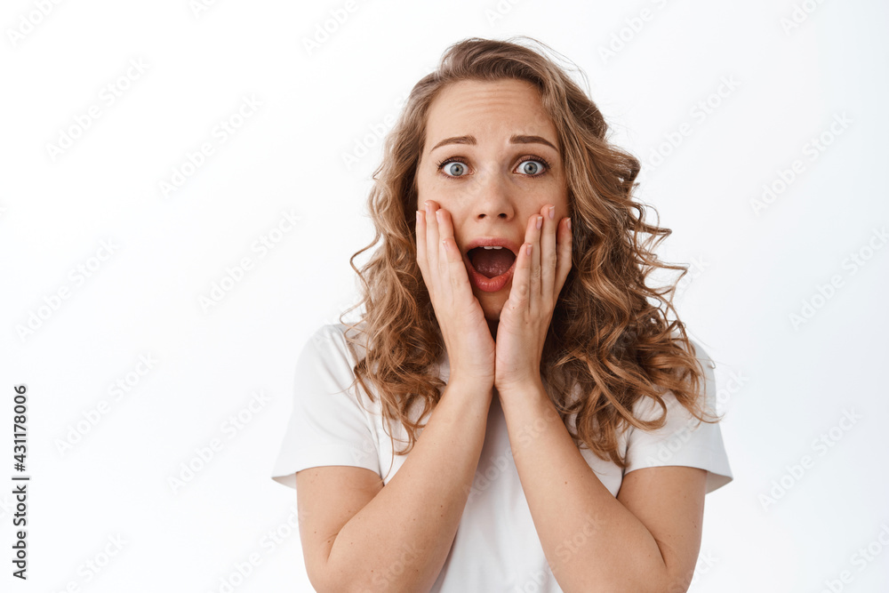 Scared and anxious blond woman screaming shocked, staring nervous and alarmed at camera, holding hands on face, panicking, standing over white background Scared and anxious blond woman screaming shocked, staring nervous and alarmed at camera, holding hands on face, panicking, standing over white background
