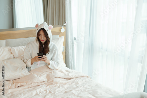 Pretty young woman in pajamas sitting on bed after waking up and checking text messages and posts on social media