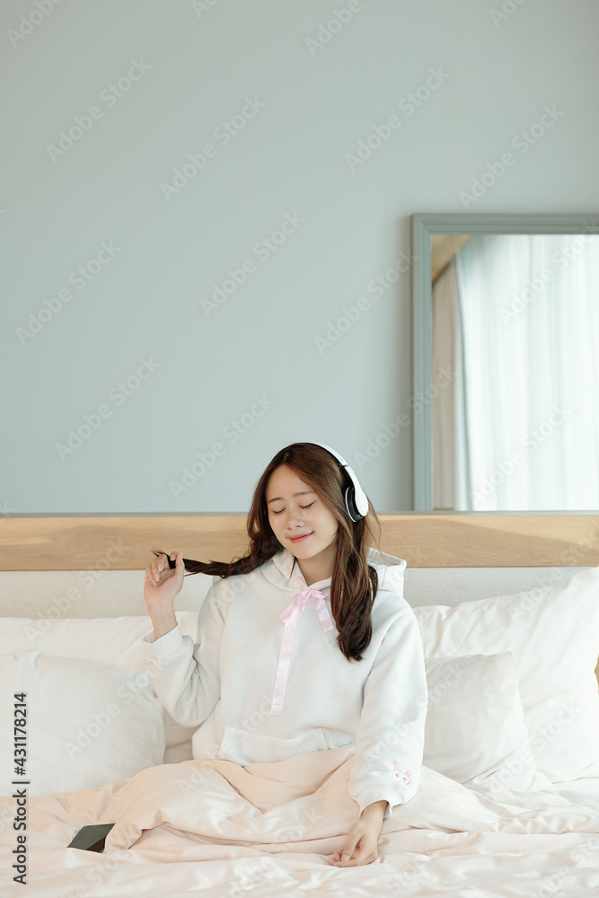 Happy beautiful young Asian woman sitting on bed and ejoying good music in her headphones Happy beautiful young Asian woman sitting on bed and ejoying good music in her headphones
