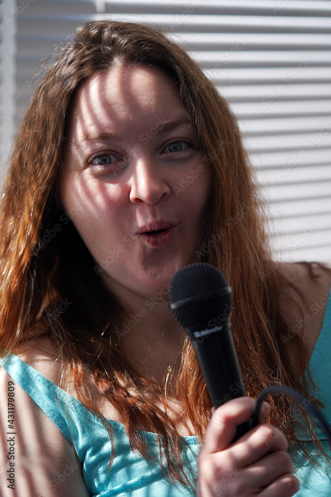 A brunette plus size girl with a microphone in her hands is standing by the window blinds. Self-isolation singing lessons and self-development. Girl in blue, woman portrait. A brunette plus size girl with a microphone in her hands is standing by the window blinds. Self-isolation singing lessons and self-development. Girl in blue, woman portrait.