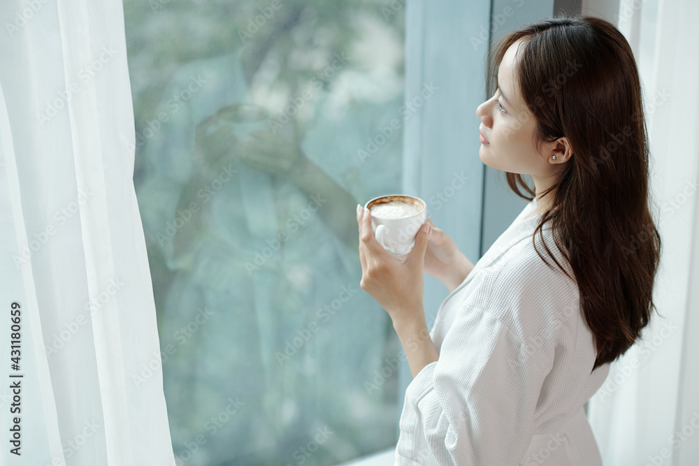 Pensive young woman drinking cup of morning coffee and looking outside through apartment window