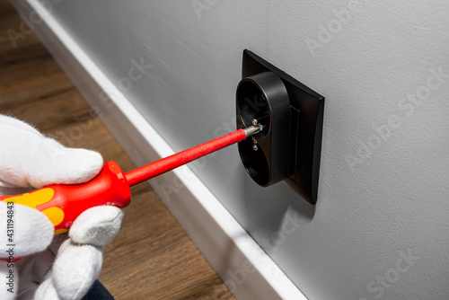 Photography An electrician uses a red screwdriver to screw a black double grounded electrical socket into the electrical box in the room wall