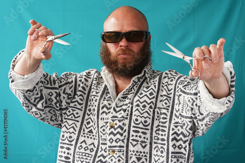 A guy with a bald head and a lush beard holds scissors and a comb in his hands. Barber hairdresser. A man with glasses and a shirt.  Stylish bearded man. Portrait                            