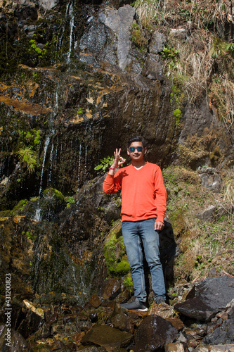 adult boy outdoor picture with waterfall and giving OK gesture wearing sunglasses and orange sweater
