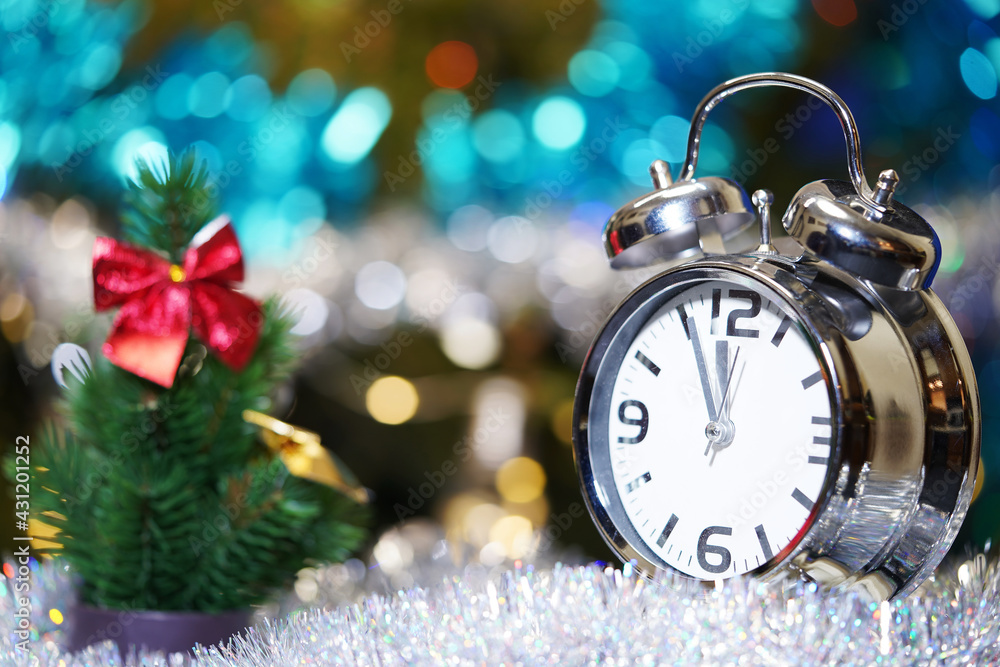 A small alarm clock shows 12 o'clock, stands on a light table with a blurred background and bokeh lights. Christmas concept. Minutes about New year.  Midnight, midday.                                 