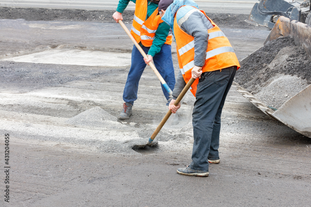 Two road workers rake old broken asphalt off the road and load it into a grader bucket. Two road workers rake old broken asphalt off the road and load it into a grader bucket.