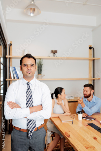 Portrait of smiling confident Indian entrepreneur crossed arms and looking at camera, his colleagues are talking at table in meeting room
