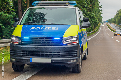 German police car on a lane and hard shoulder of a motorway. switched on headlights and flasher on the police car. Concrete asphalt with two lanes. Rainy weather in spring with green trees