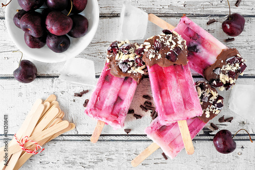 Summer chocolate and almond dipped cherry popsicles. Above view table scene on a rustic white wood background.