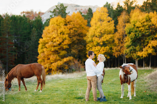 Man hugs a woman while standing on the lawn in the autumn forest. Horses grazing on the lawn