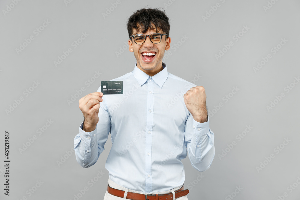 Young fun rich smiling employee business latin man corporate lawyer 20s wearing classic white shirt glasses hold in hand credit bank card do winner gesture isolated on grey background studio portrait Young fun rich smiling employee business latin man corporate lawyer 20s wearing classic white shirt glasses hold in hand credit bank card do winner gesture isolated on grey background studio portrait