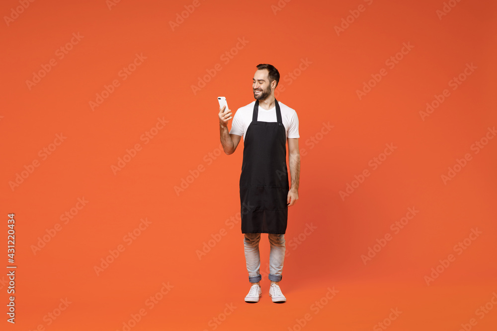 Full length young happy fun man barista bartender barman employee in black apron white tshirt work in coffee shop using mobile cell phone isolated on orange background. Small business startup concept Full length young happy fun man barista bartender barman employee in black apron white tshirt work in coffee shop using mobile cell phone isolated on orange background. Small business startup concept
