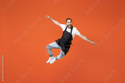Full length young man barista bartender barman employee in black apron white tshirt work in coffee shop jump high outstretched hands legs isolated on orange background. Small business startup concept
