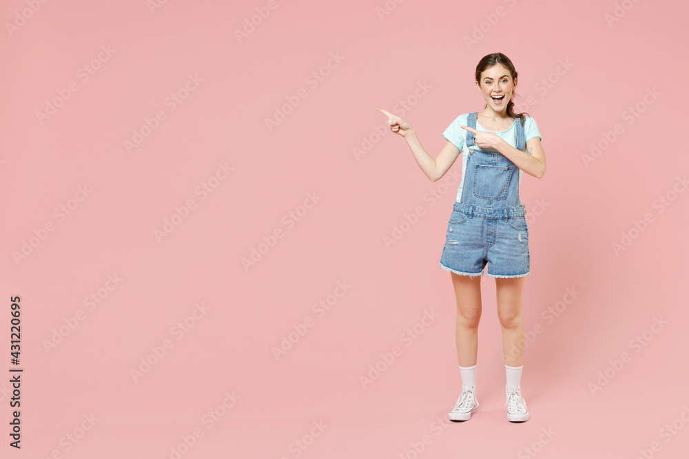 Full length young smiling happy cheerful caucasian woman 20s in trendy denim clothes blue t-shirt point index finger aside on workspace area mock up isolated on pastel pink background studio portrait Full length young smiling happy cheerful caucasian woman 20s in trendy denim clothes blue t-shirt point index finger aside on workspace area mock up isolated on pastel pink background studio portrait