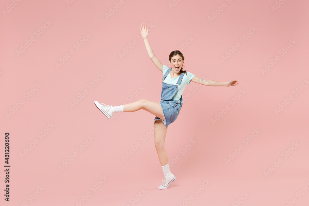 Full length young smiling joyful caucasian woman 20s in trendy denim clothes blue tshirt stand on toes dancing with outstretched hands raised up leg isolated on pastel pink background studio portrait