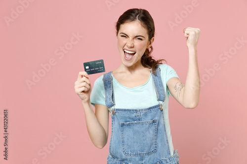 Young happy caucasian woman 20s in trendy denim clothes blue t-shirt pointing index finger on credit bank card do winner gesture clench fist isolated on pastel pink color background studio portrait