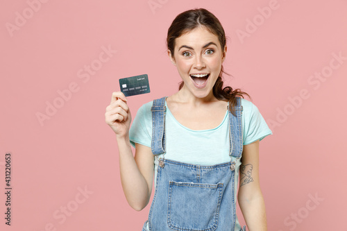 Young overjoyed positive excited happy caucasian woman 20s wear trendy denim clothes blue t-shirt hold credit bank card isolated on pastel pink background studio portrait. People lifestyle concept