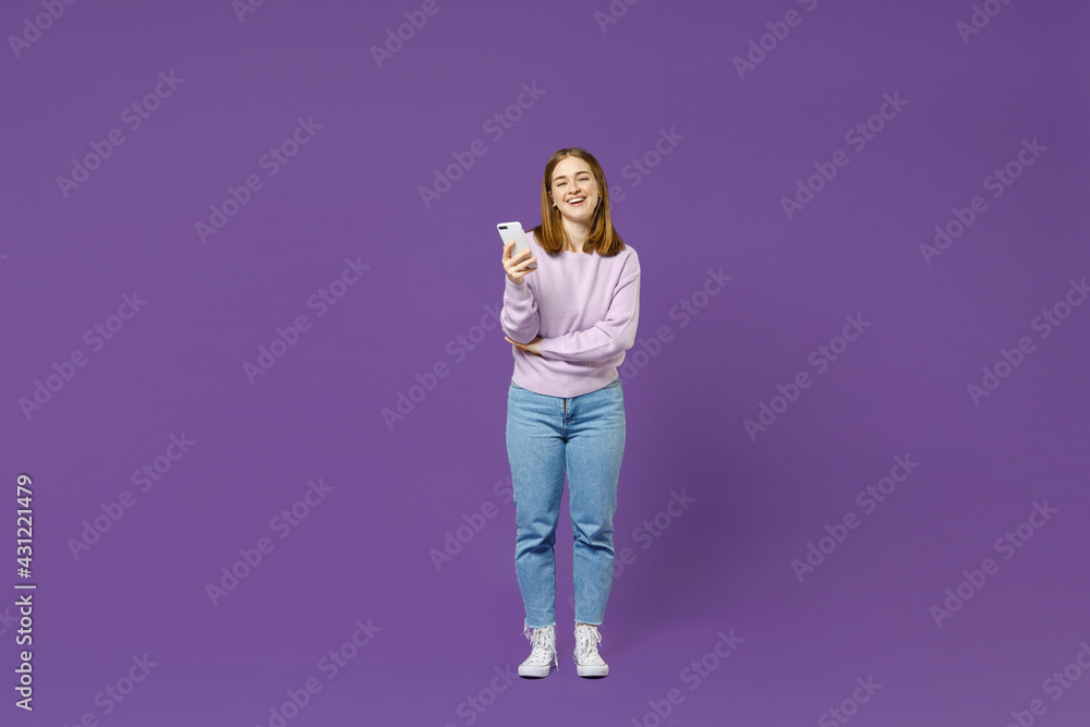 Full length young smiling positive happy cheerful student caucasian woman 20s in purple knitted sweater using mobile cell phone isolated on violet background studio portrait. People lifestyle concept. Full length young smiling positive happy cheerful student caucasian woman 20s in purple knitted sweater using mobile cell phone isolated on violet background studio portrait. People lifestyle concept.