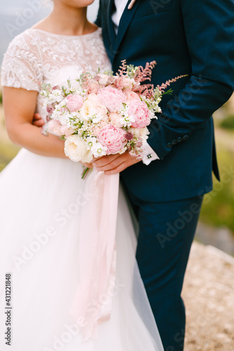 The bride and groom stand hugging and hold the bride's bouquet with delicate pink roses, peonies and astilbe, close-up 