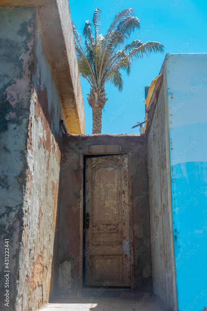 A fragment of an old Egyptian house on the beach. A wooden door to a one-story abandoned house, a palm tree and a blue sky.