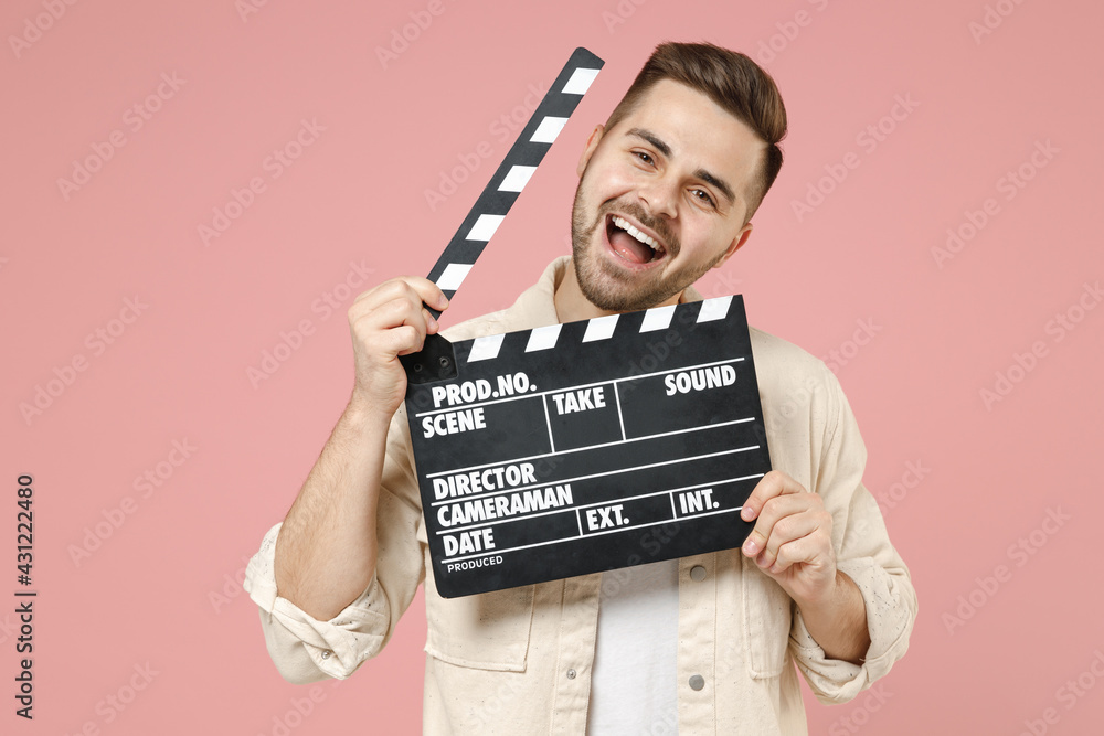 Young smiling laughing cheerful fun man wear jacket white t-shirt holding classic black film making clapperboard isolated on pastel pink color background studio portrait. Lifestyle people concept.