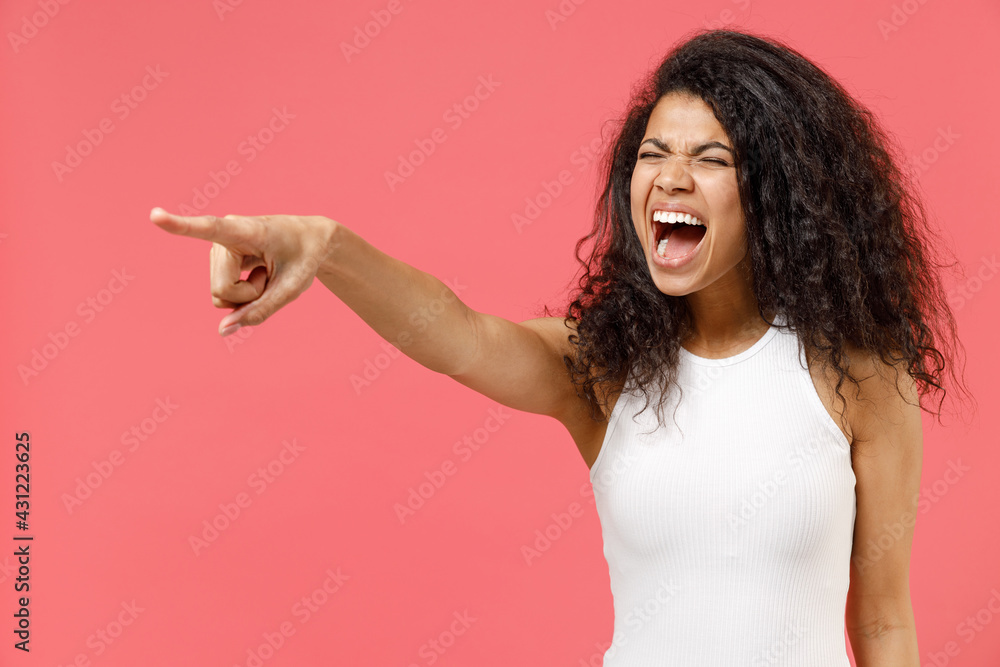 Young indignant strict sad angry african american woman 20s wearing casual white tank shirt point index finger aside scream shout command do it isolated on pink background. People lifestyle concept. Young indignant strict sad angry african american woman 20s wearing casual white tank shirt point index finger aside scream shout command do it isolated on pink background. People lifestyle concept.