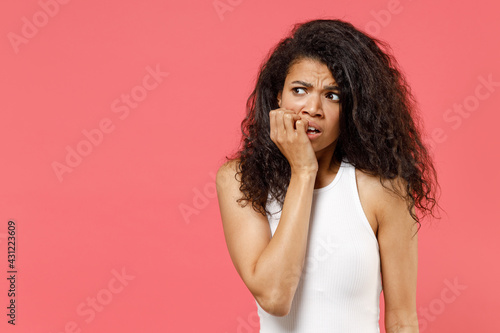 Young puzzled thoughtful pensive sad confused troubled african american woman in casual white tank shirt look aside biting nails fingers think isolated on pink background. People lifestyle concept