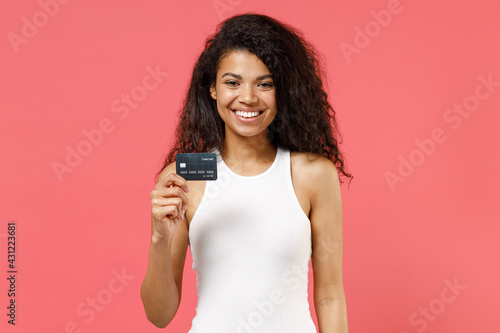 Young fun smiling happy cheerful successful satisfied positive african american woman 20s wear casual white tank shirt hold in hand credit bank card isolated on pink color background studio portrait.
