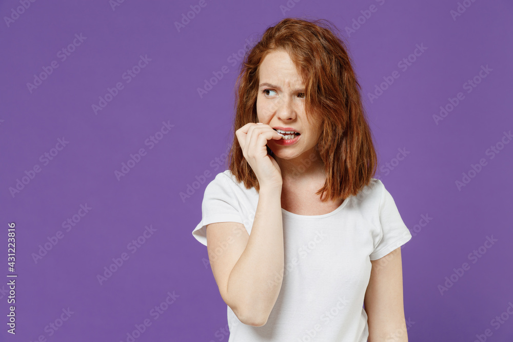 Young sad puzzled troubled pensive thoughtful confused caucasian woman 20s wearing white basic t-shirt looking aside, biting nails, fingers isolated on dark violet color background studio portrait Young sad puzzled troubled pensive thoughtful confused caucasian woman 20s wearing white basic t-shirt looking aside, biting nails, fingers isolated on dark violet color background studio portrait
