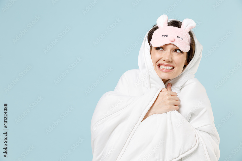 Young woman in pajamas jam sleep eye mask rest relaxing at home wrap under blanket duvet cover ear neighbors noise snoring isolated on pastel blue background studio. Bad mood night bedtime concept.