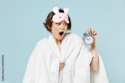 Young shock confused woman in pajamas jam sleep eye mask rest relaxing at home wrap cover under blanket duvet hold clock alarm isolated on pastel blue background studio Bad mood night bedtime concept. photo