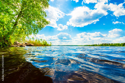 Forest lake with trees growing on the shore. Close up view from the water level. Waves on the lake. Cumulus clouds. Sunlight illuminates the clouds. Beautiful nature. Russia, Europe.