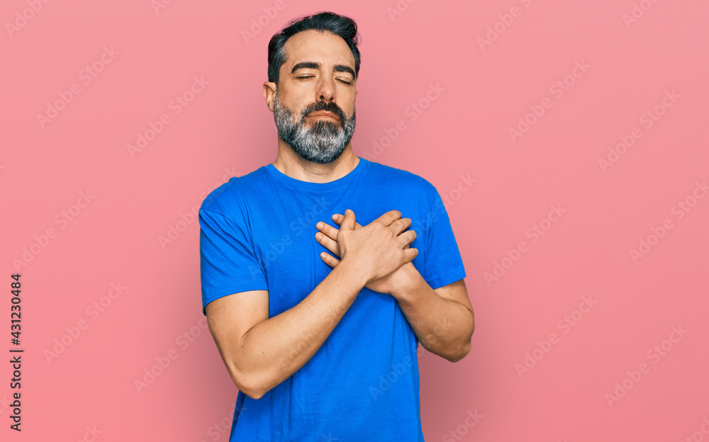 Middle aged man with beard wearing casual blue t shirt smiling with hands on chest with closed eyes and grateful gesture on face. health concept. Middle aged man with beard wearing casual blue t shirt smiling with hands on chest with closed eyes and grateful gesture on face. health concept.