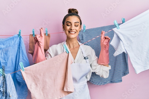 Beautiful brunette young woman washing clothes at clothesline gesturing finger crossed smiling with hope and eyes closed. luck and superstitious concept.