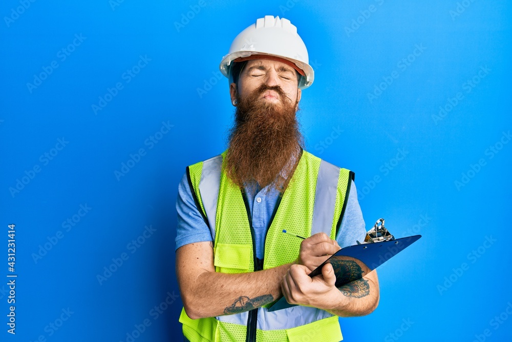 Fototapeta premium Redhead man with long beard wearing safety helmet holding clipboard looking at the camera blowing a kiss being lovely and sexy. love expression.