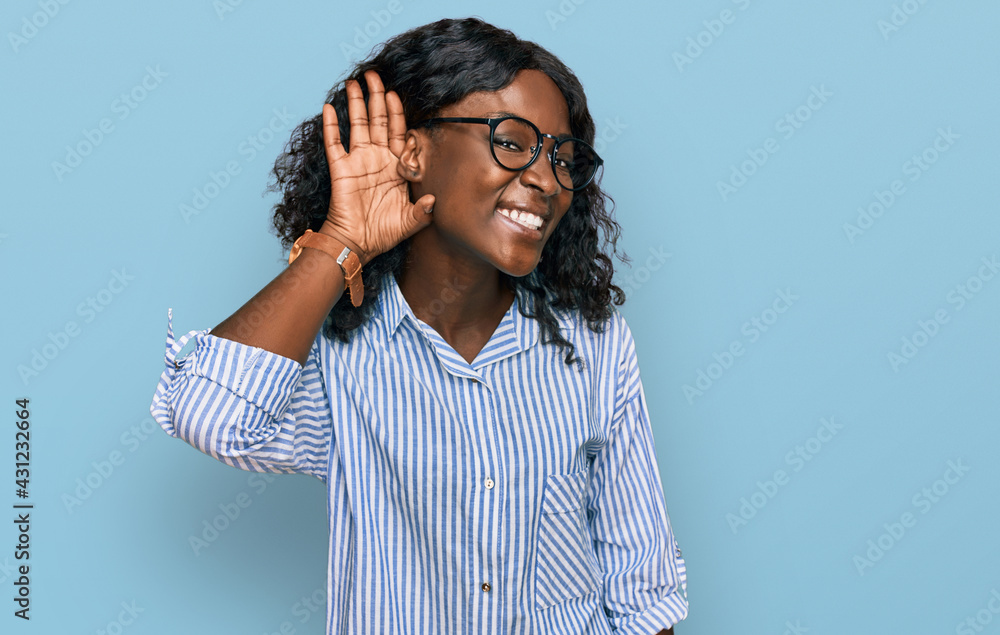 Beautiful african young woman wearing casual clothes and glasses smiling with hand over ear listening an hearing to rumor or gossip. deafness concept. Beautiful african young woman wearing casual clothes and glasses smiling with hand over ear listening an hearing to rumor or gossip. deafness concept.