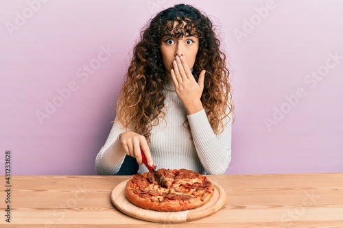 Young hispanic girl cutting tasty pepperoni pizza covering mouth with hand  shocked and afraid for mistake. surprised expression