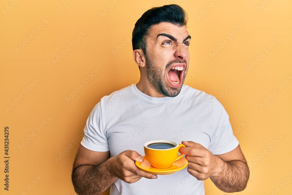 Young hispanic man drinking a cup of coffee angry and mad screaming frustrated and furious, shouting with anger looking up. Young hispanic man drinking a cup of coffee angry and mad screaming frustrated and furious, shouting with anger looking up.