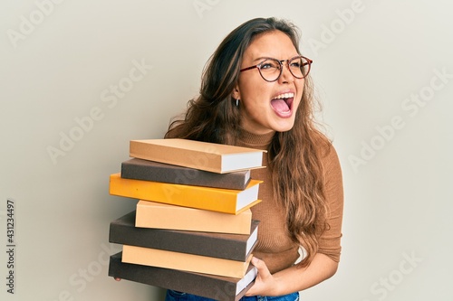Young hispanic girl holding a pile of books angry and mad screaming frustrated and furious, shouting with anger. rage and aggressive concept.