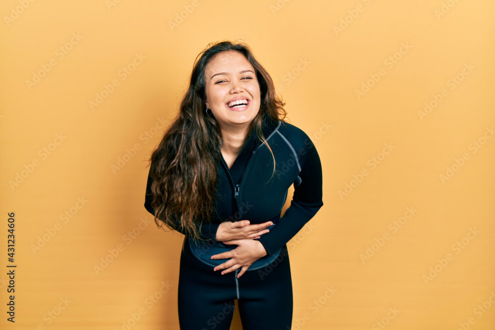 Young hispanic girl wearing diver neoprene uniform smiling and laughing hard out loud because funny crazy joke with hands on body. Young hispanic girl wearing diver neoprene uniform smiling and laughing hard out loud because funny crazy joke with hands on body.