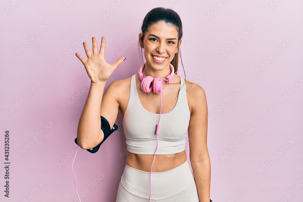 Young hispanic woman wearing gym clothes and using headphones showing and pointing up with fingers number five while smiling confident and happy.