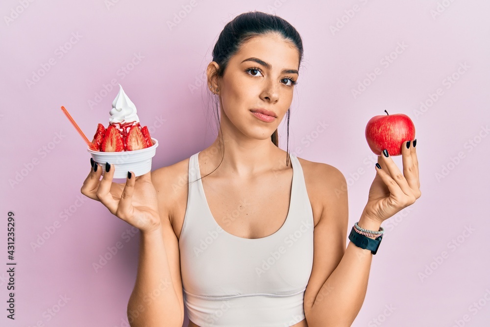 Young hispanic woman wearing sportswear holding apple and ice cream relaxed with serious expression on face. simple and natural looking at the camera. Young hispanic woman wearing sportswear holding apple and ice cream relaxed with serious expression on face. simple and natural looking at the camera.