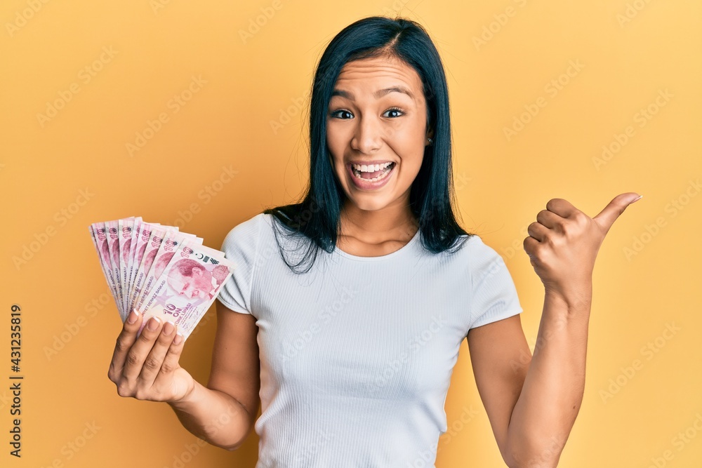 Beautiful hispanic woman holding 10 turkish lira banknotes pointing thumb up to the side smiling happy with open mouth Beautiful hispanic woman holding 10 turkish lira banknotes pointing thumb up to the side smiling happy with open mouth