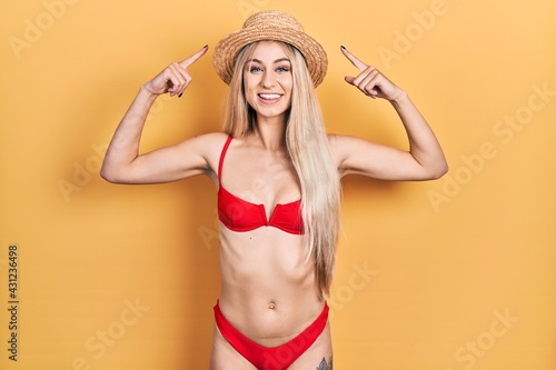 Young caucasian woman wearing bikini and summer hat smiling pointing to head with both hands finger, great idea or thought, good memory