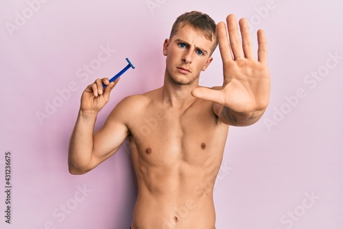 Young caucasian man shirtless holding razor with open hand doing stop sign with serious and confident expression, defense gesture